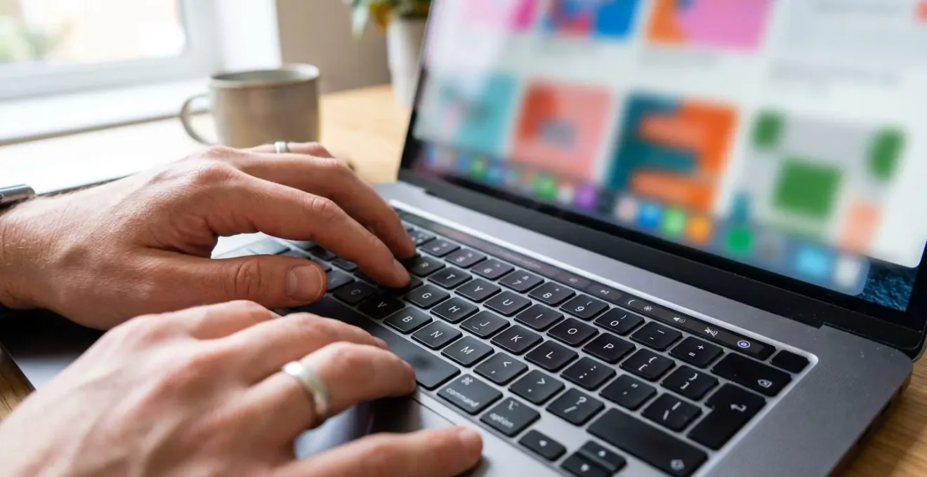 Close-up view of hands typing on modern laptop keyboard with blurred design software interface showing colour palette in soft background bokeh